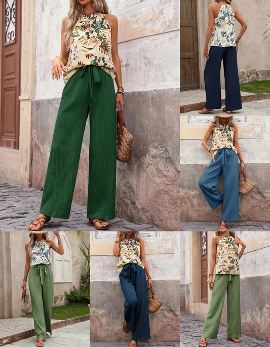 Woman wearing a floral top and wide-leg pants in green and blue, standing against a stone wall.